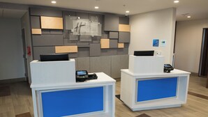 A reception desk with a digital screen and a cash register, set against a wall with a geometric design and a sign indicating a Holiday Inn Express. - Holiday Inn Express Hotel & Suites East Lansing by IHG (East Lansing)