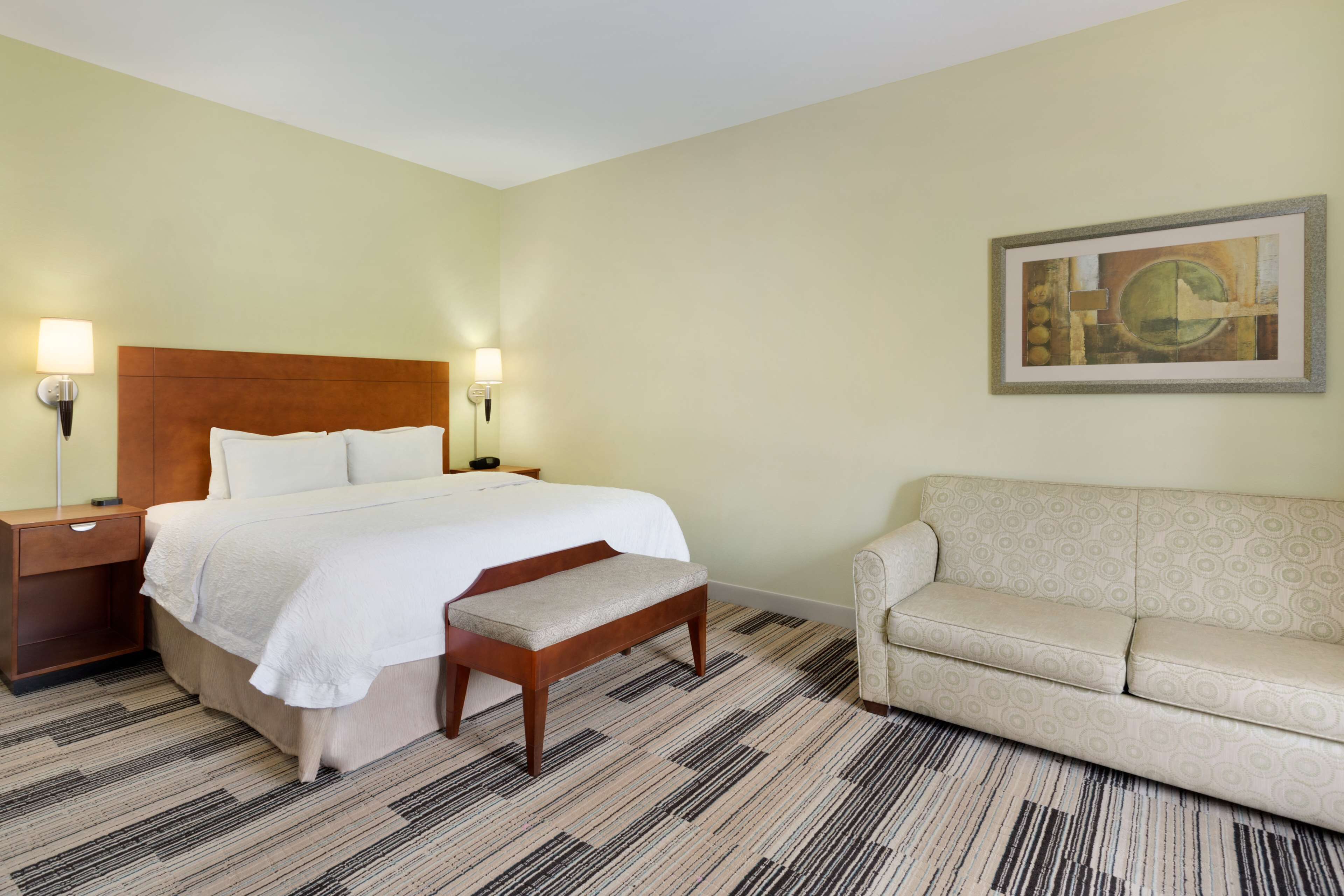 A hotel room with a bed, bedside tables, a bench, a sofa, and a painting on the wall.