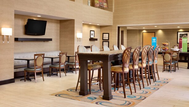 A hotel dining area with a large table and chairs, a TV mounted on the wall, and a buffet counter in the background. - Hampton Inn & Suites Birmingham East Irondale (Birmingham)