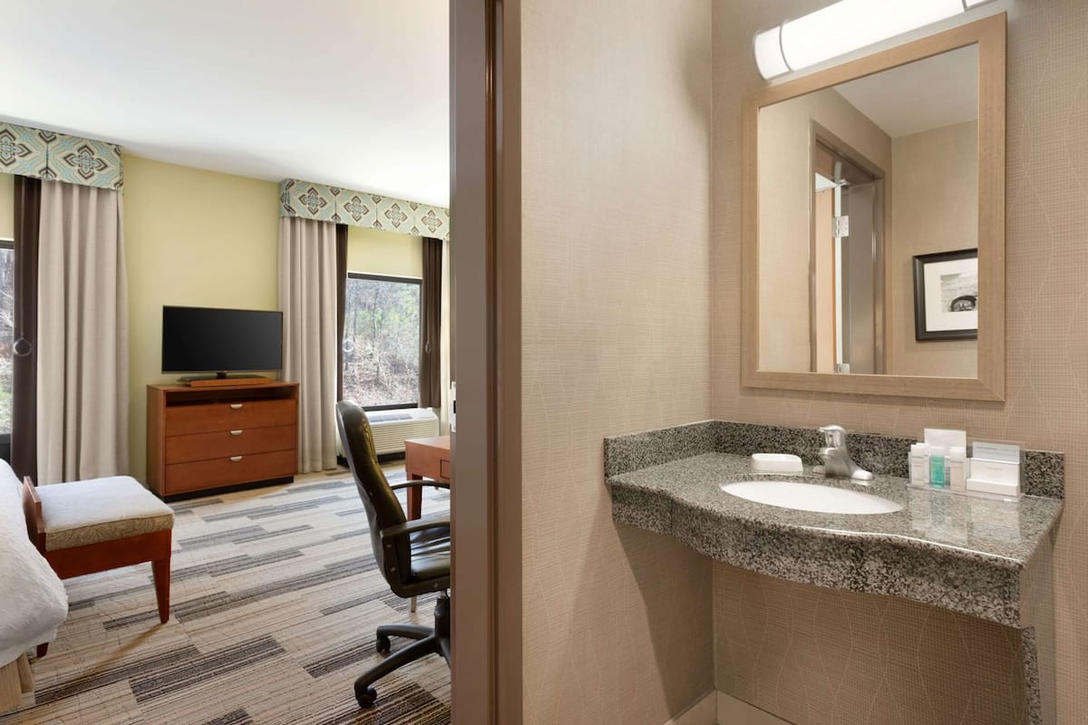 A hotel room with a bed, a TV on a wooden dresser, a desk with a chair, a bathroom with a granite countertop sink, and a large mirror.