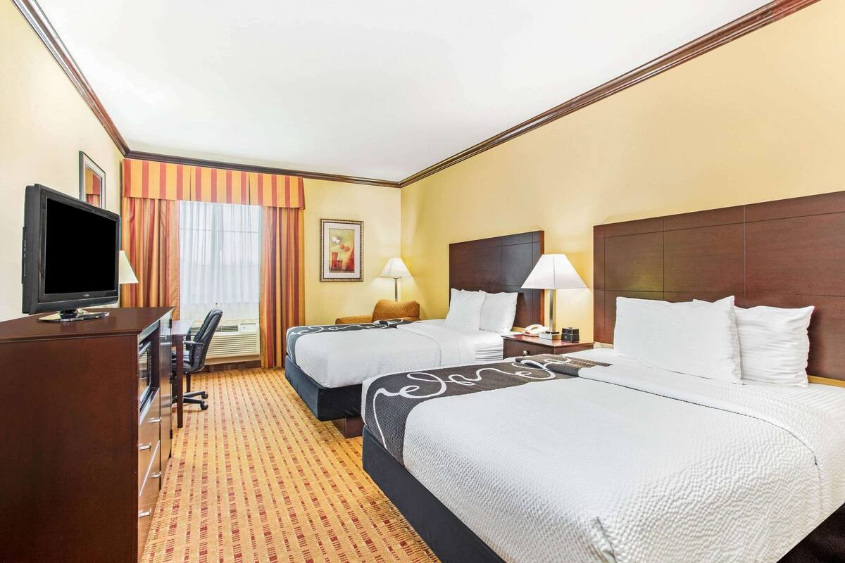 Ein Hotelzimmer mit zwei Betten, einem Schreibtisch, einem Fernseher, einer Lampe und einem Fenster mit Vorhängen.