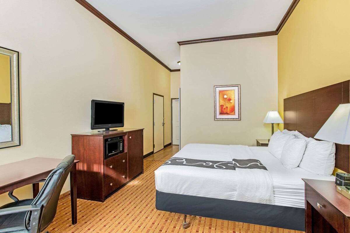 Ein Hotelzimmer mit einem Bett, einem Schreibtisch mit Fernseher, einem Sessel, einer Lampe und einem gerahmten Bild an der Wand.