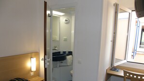 A hotel room with a bed, a bedside table with a lamp, a wooden chair, a desk with a mirror, and a bathroom visible through an open door. - Mister Bed Lomme (Lille)