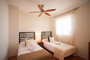 A hotel room with two single beds, a ceiling fan, a nightstand with a lamp, and a window with sheer curtains. - Apartamentos Paraiso Playa (Vera)