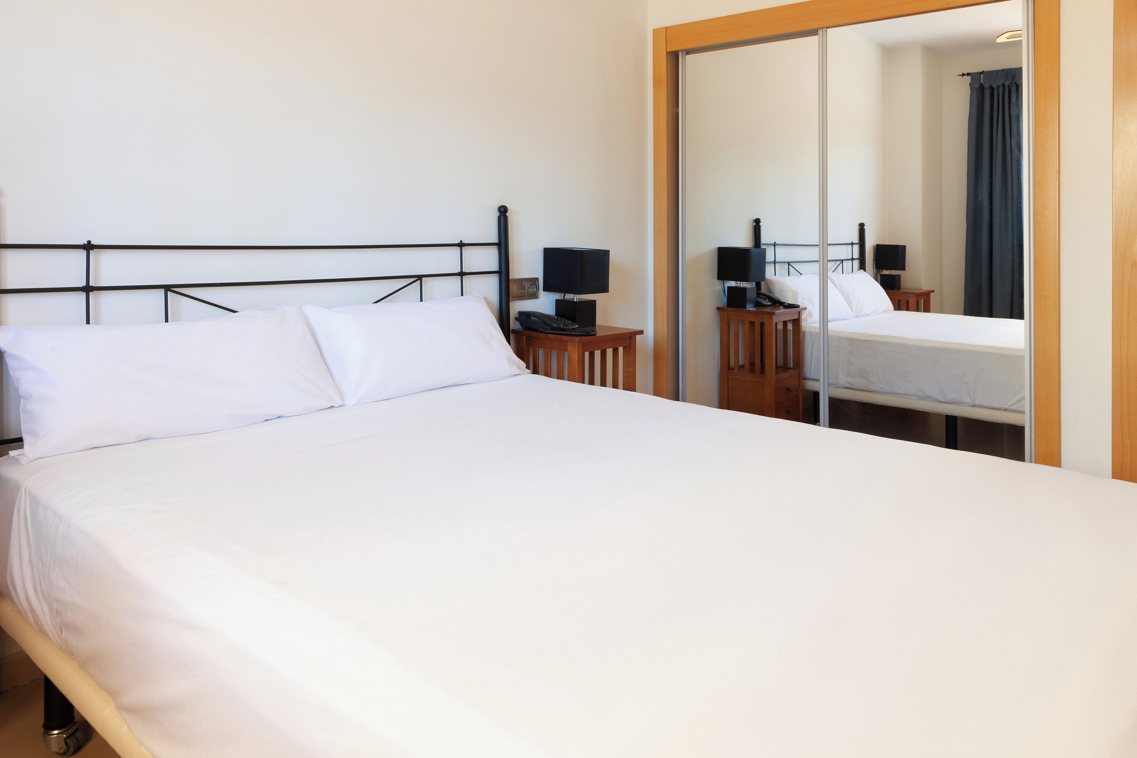 A neatly made bed with white linens, a bedside table with a lamp, and a mirror reflecting the room.