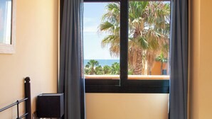 A hotel room with a bed, a desk, and a view of palm trees and the ocean through a window. - Apartamentos Playamarina (Ayamonte)