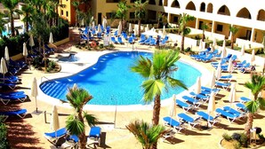 A pool area with blue lounge chairs and palm trees, surrounded by a building with arches and a view of the sea. - Apartamentos Playamarina (Ayamonte)