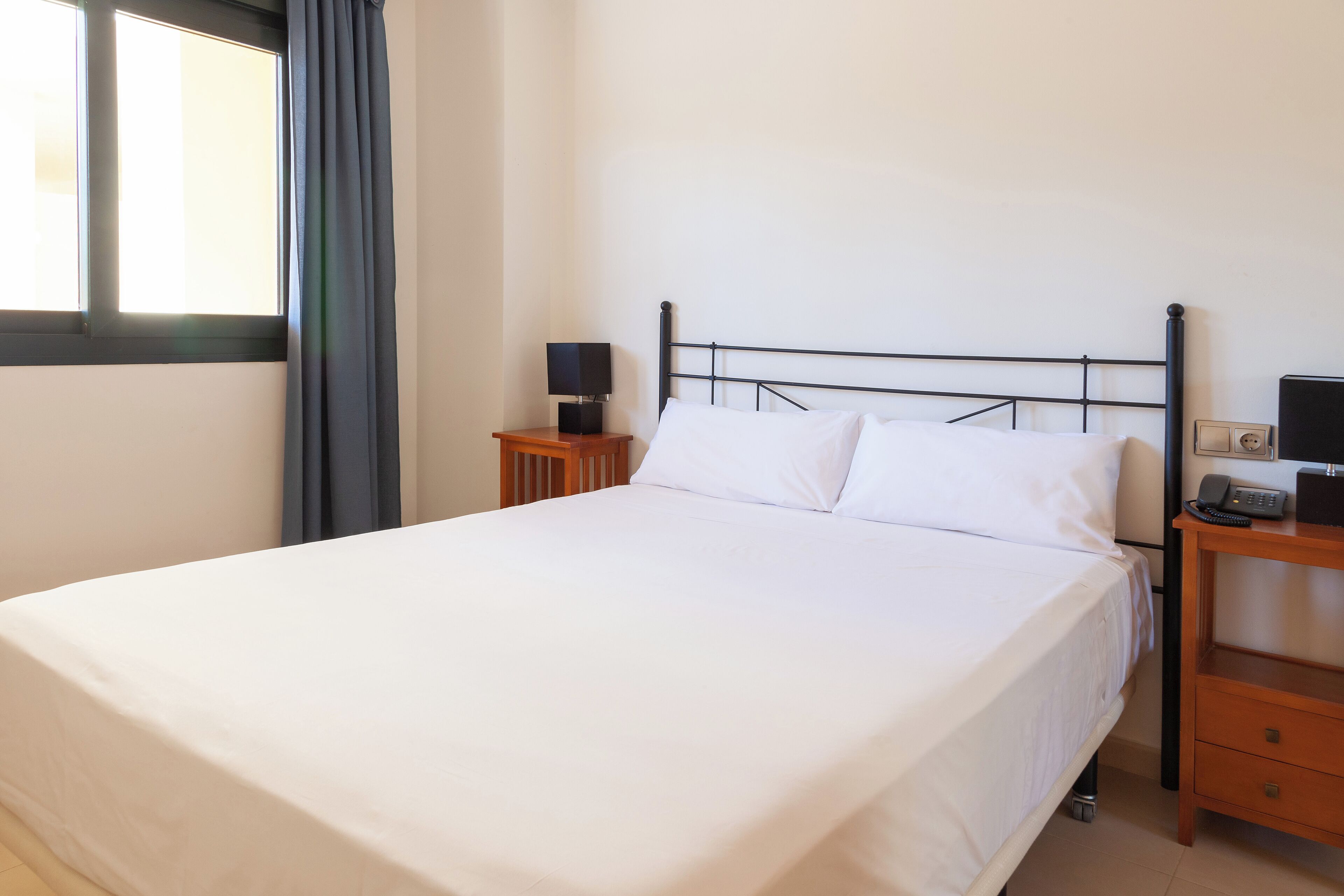 A neatly made bed with white linens, a bedside table with a lamp, and a small bedside table with a phone.
