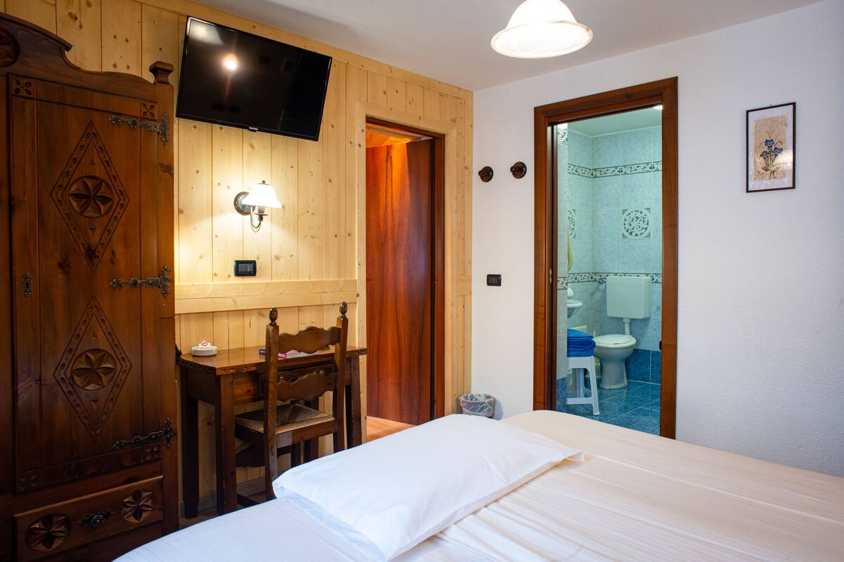 A wooden room with a bed, a desk, a chair, a TV mounted on the wall, and a bathroom visible through an open door.