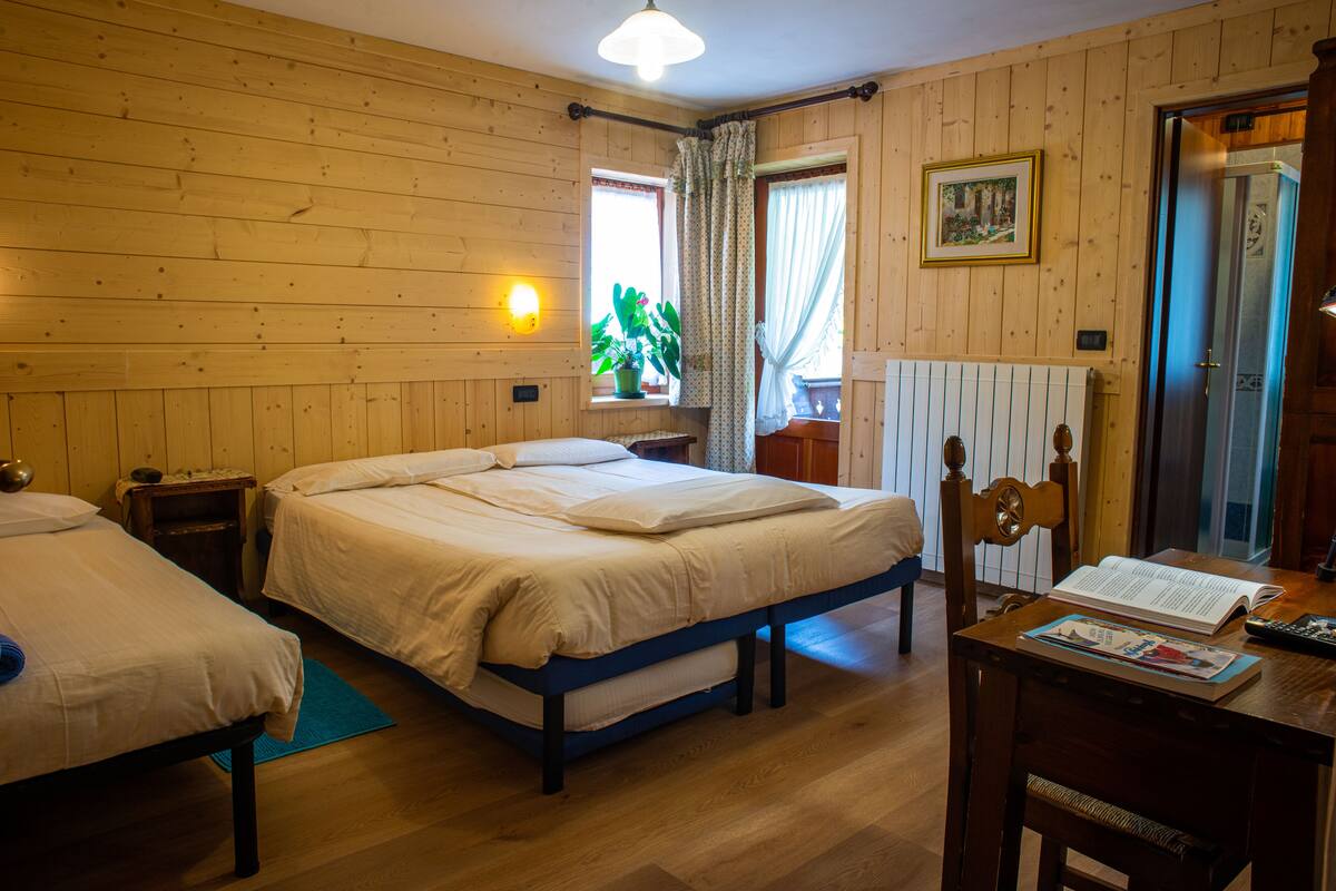 A wooden-paneled room with two beds, a table with a book and a remote control, a window with curtains, and a radiator.