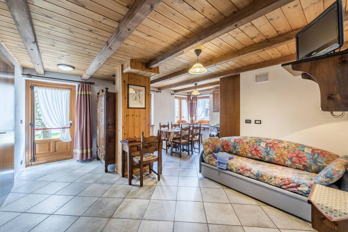 A cozy living room with wooden beams, a sofa, a dining table with chairs, and a television mounted on the wall.