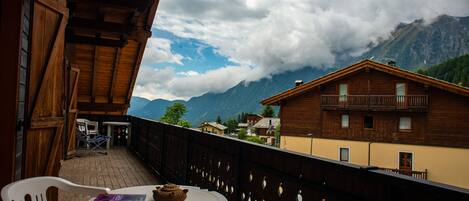 A wooden balcony with a table and chairs, overlooking a mountain village with houses and a view of the mountains.
