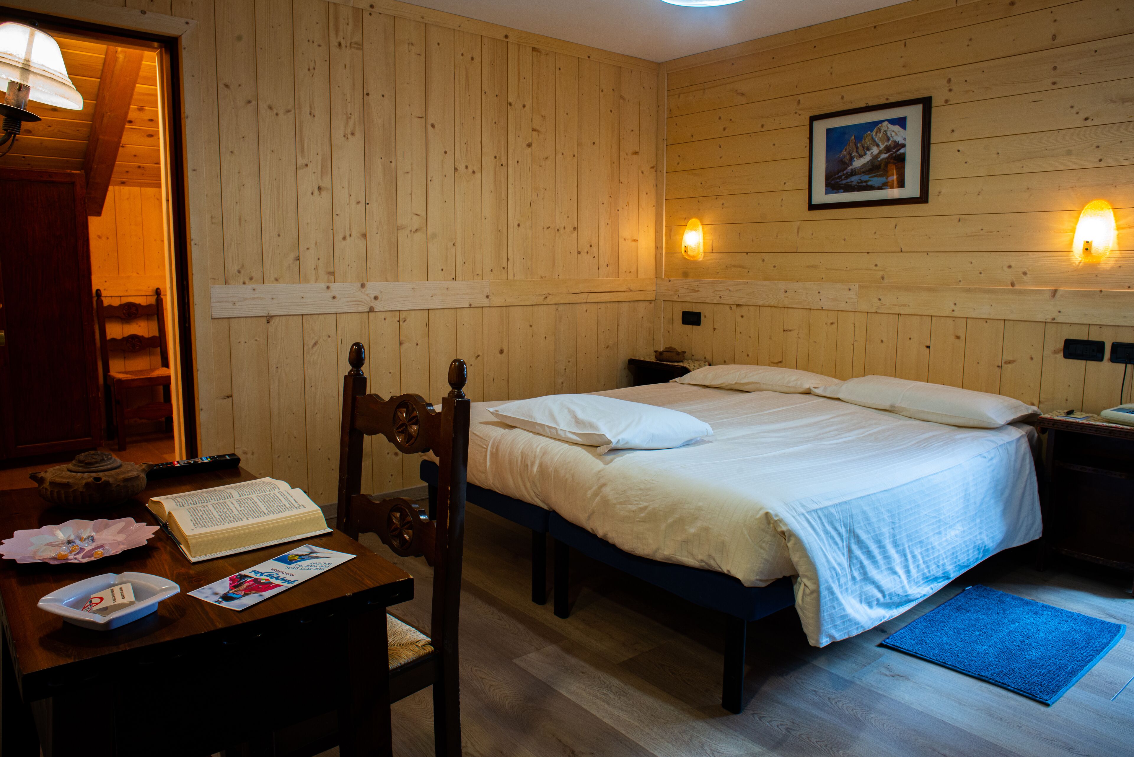 Ein Zimmer mit Holzwänden, einem Bett, einem Schreibtisch mit einem Buch, einem Stuhl und einem gerahmten Bild an der Wand.