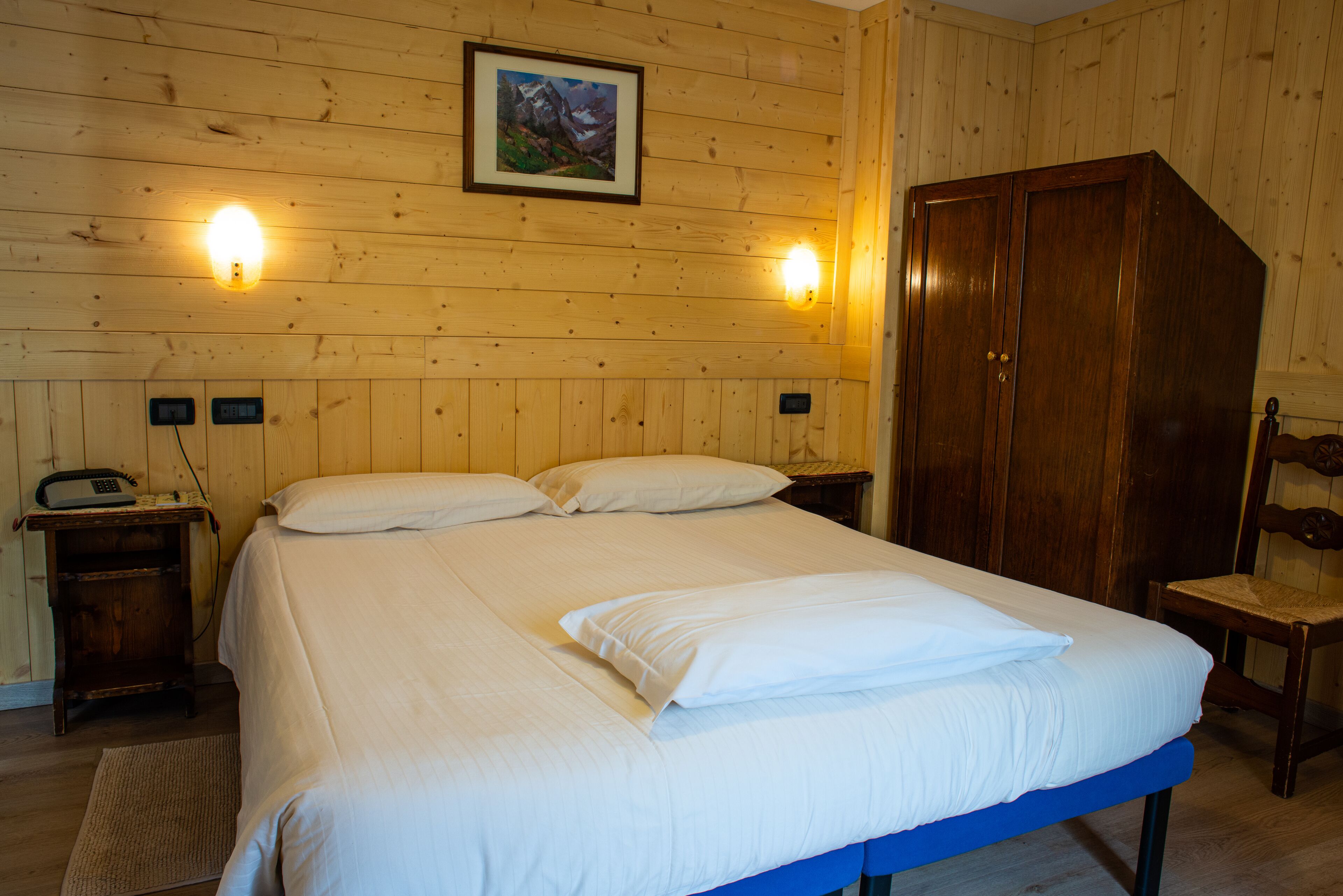 A wooden-paneled room with a bed, two bedside lamps, a wooden wardrobe, and a chair.