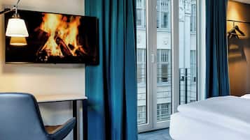 A hotel room with a TV displaying a fireplace, a desk, a chair, a bed, and a view of the city through the window.