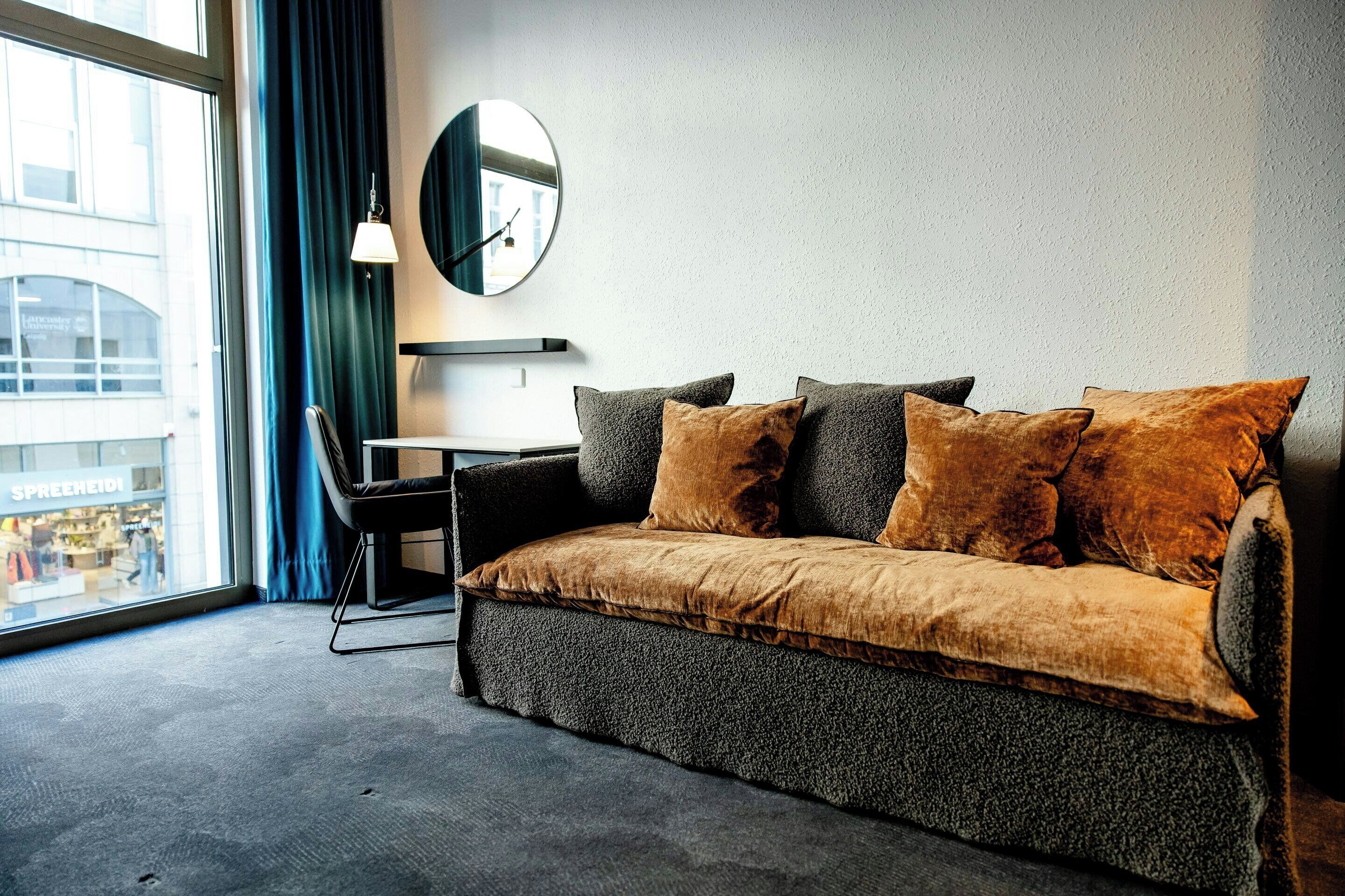 Habitación de hotel moderna con sofá, escritorio, espejo y vistas a un paisaje urbano.