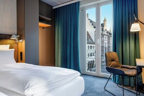 A hotel room with a bed, a chair, a desk, and a view of a historic building through the window. - Motel One Leipzig-Nikolaikirche (Leipzig)
