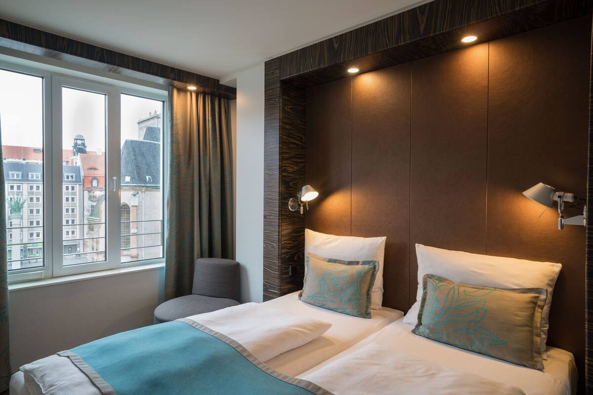 A hotel room with a large bed, a chair, a window with curtains, and a view of buildings outside.