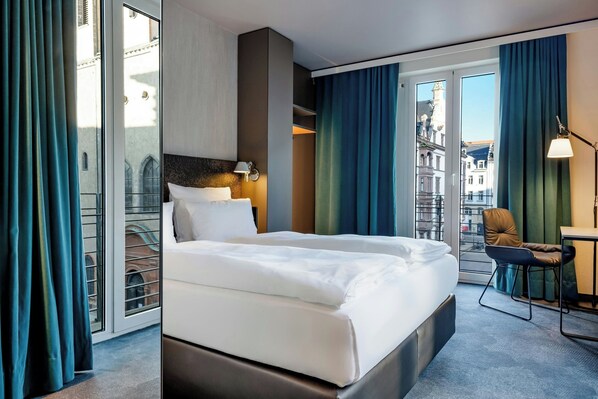 A hotel room with a large bed, a desk, a chair, and a view of the city through large windows. - Motel One Leipzig-Nikolaikirche (Leipzig)