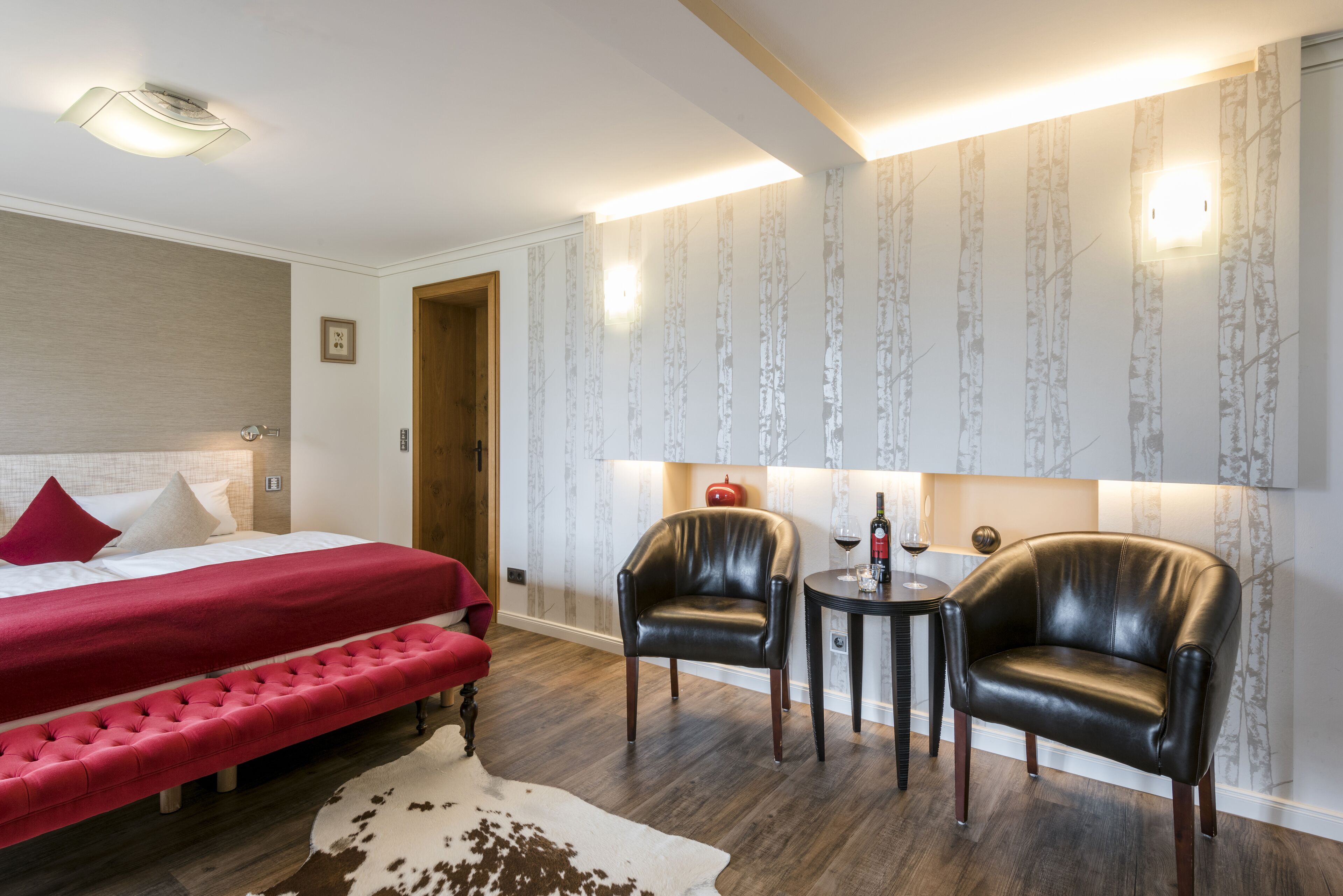 A hotel room with a bed, two leather armchairs, a small table, and a red bench.