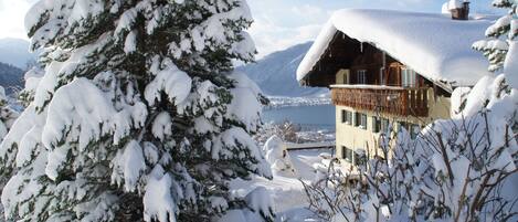 A snow-covered chalet with a balcony overlooking a lake and mountains in the background.