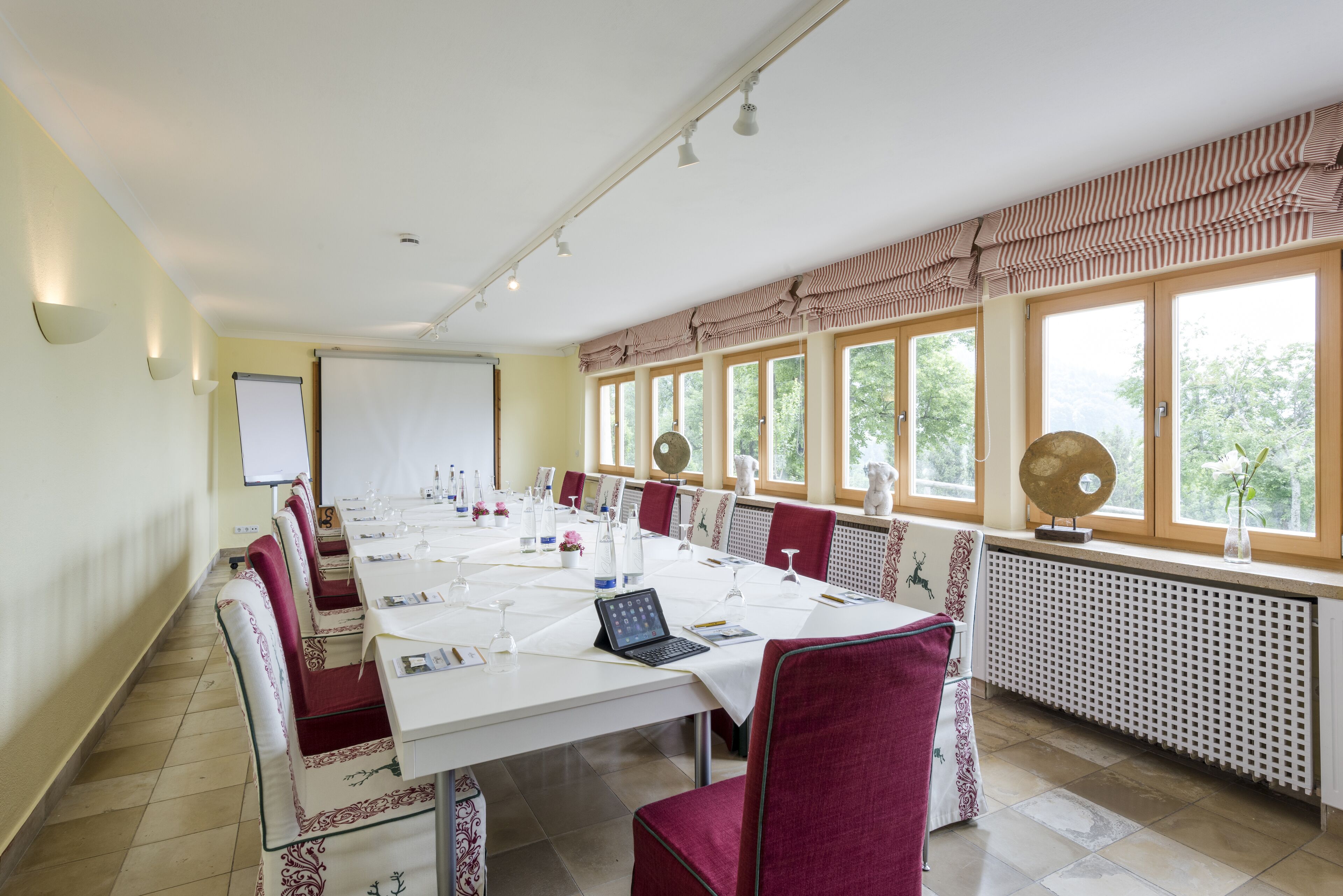 A conference room with a long table, red chairs, a whiteboard, and large windows with views of trees.