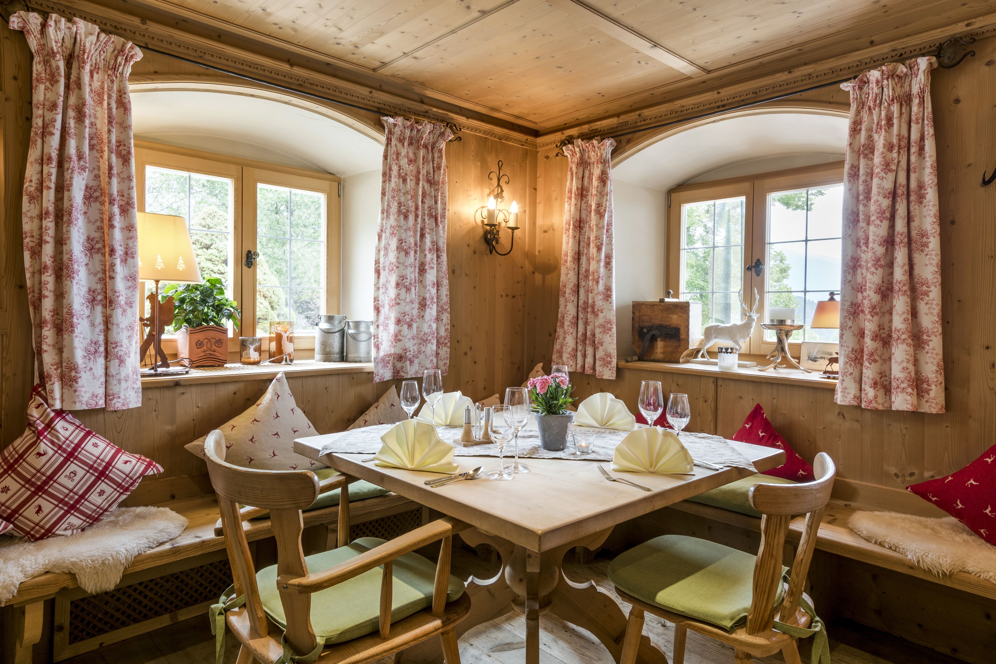 A wooden dining room with a table set for four, flanked by two windows with curtains.