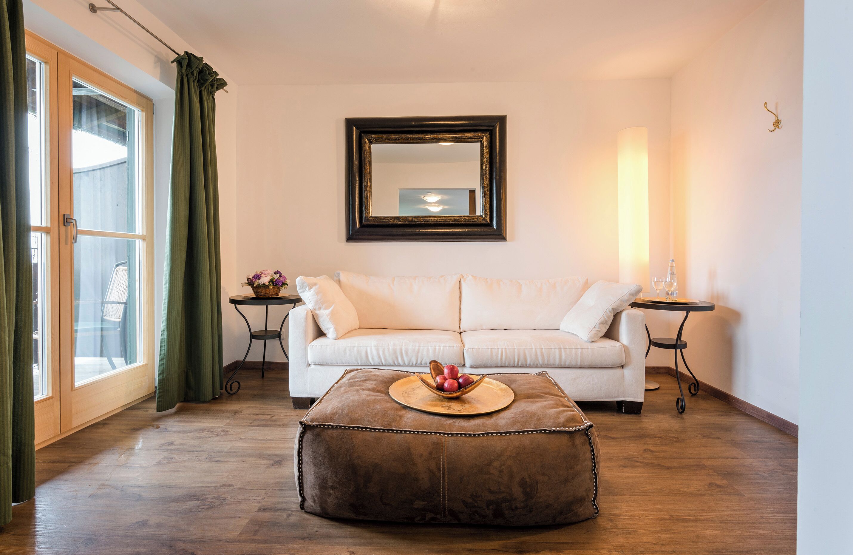 Une pièce avec un canapé blanc, un pouf marron sur lequel sont posés des fruits, une petite table et un miroir au mur.