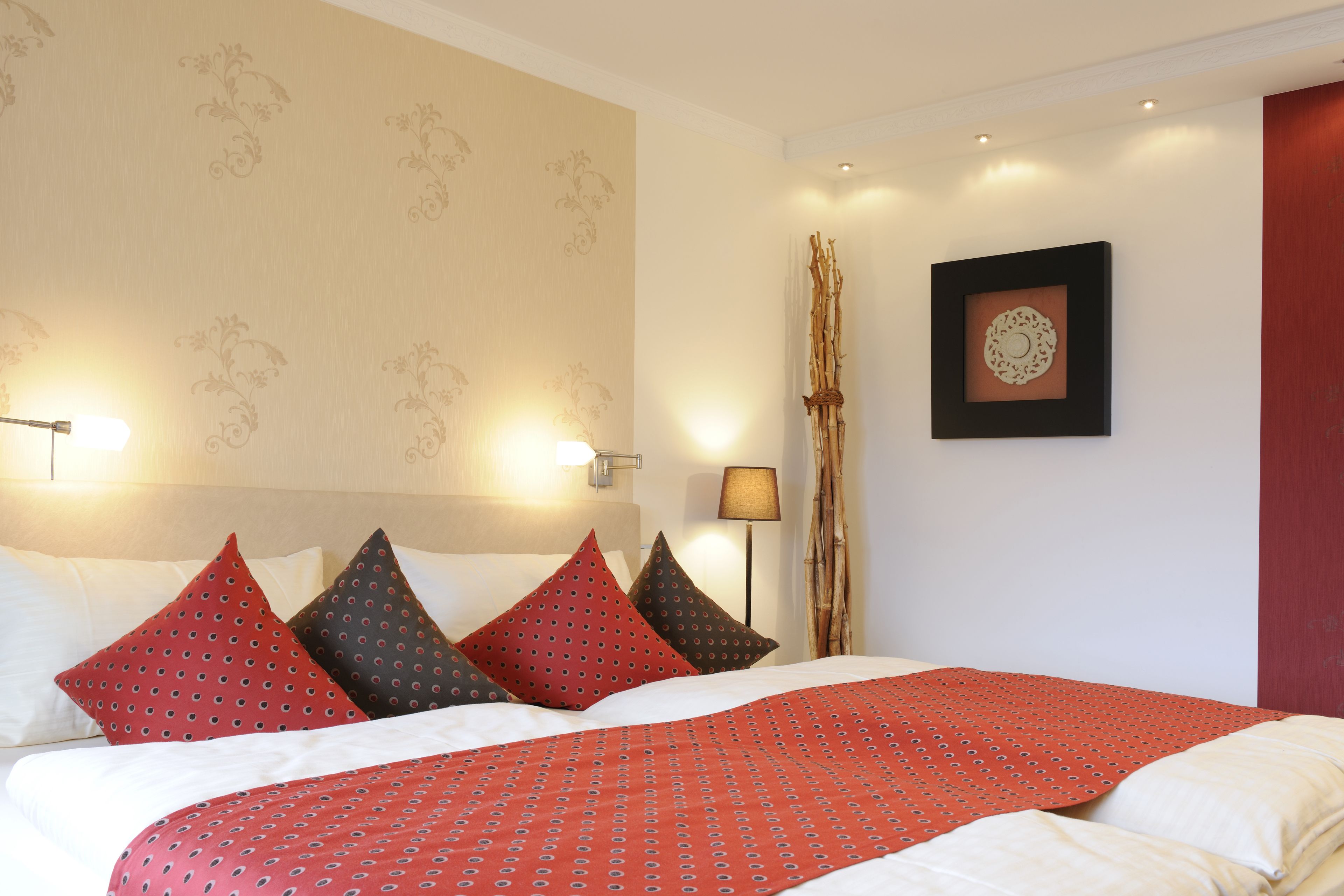 Ein ordentlich bezogenes Bett mit einer rot gemusterten Tagesdecke und mehreren Kissen, eine an der Wand befestigte Lampe und ein gerahmtes Bild.