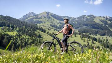 Uma pessoa ao lado de uma bicicleta de montanha numa colina gramada, com uma paisagem montanhosa ao fundo.