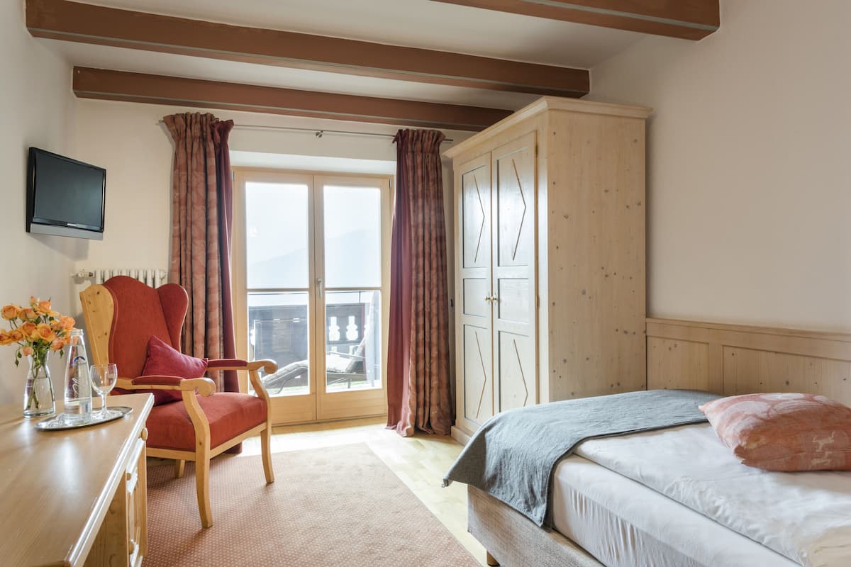 Une chambre à coucher avec un lit, une armoire en bois, un fauteuil rouge, un bureau avec un vase de fleurs et un téléviseur fixé au mur.