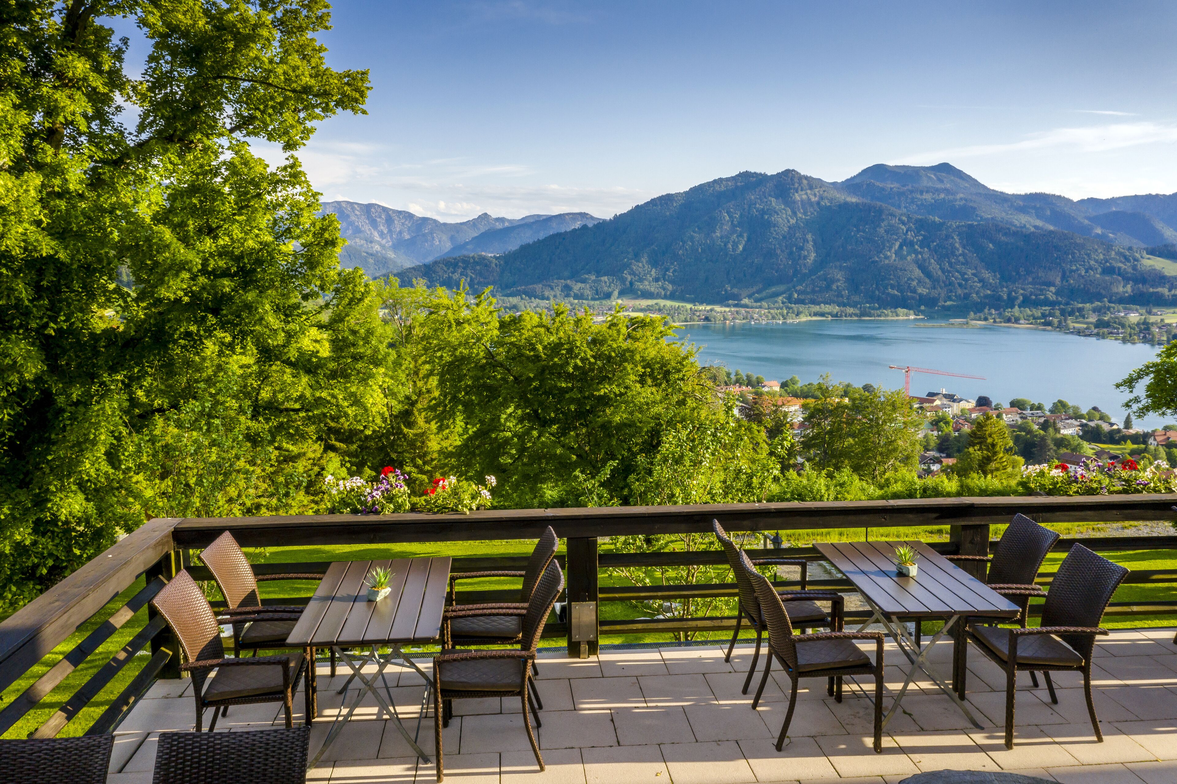 An outdoor patio with wooden tables and wicker chairs, overlooking a lake and mountains.
