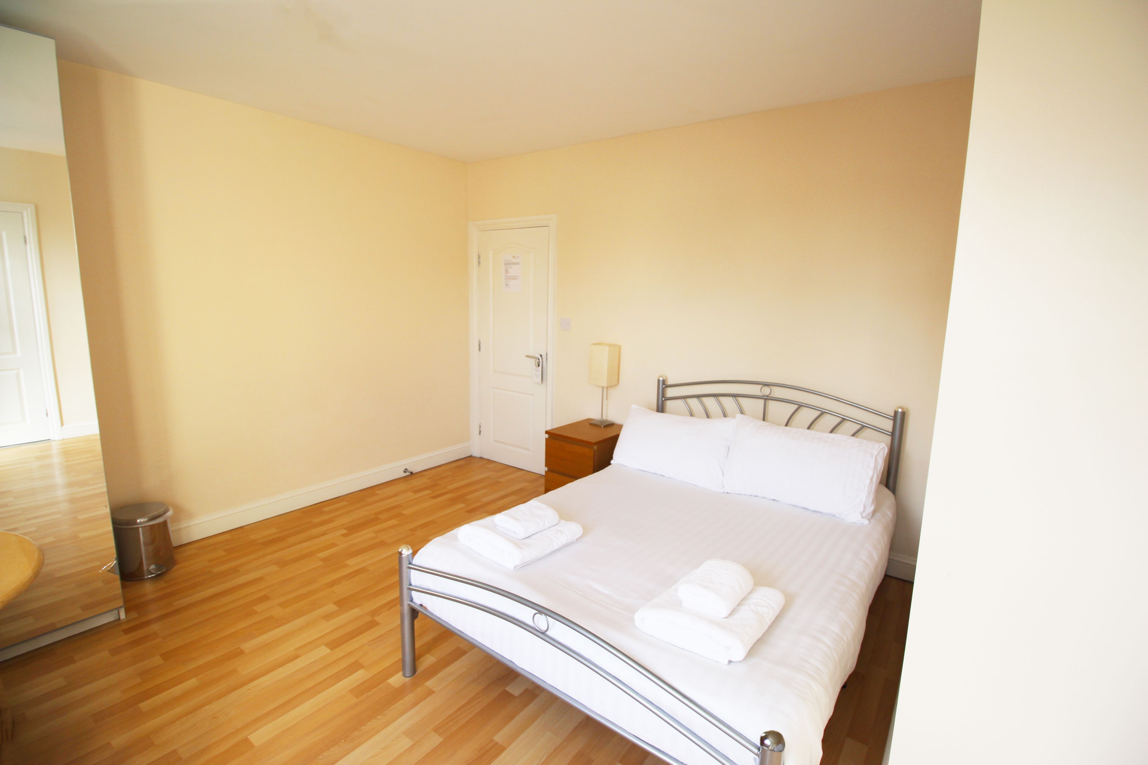 Un dormitorio con estructura de cama metálica, ropa de cama blanca, suelo de madera, mesita de noche con lámpara y una puerta.