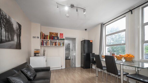 Una sala de estar moderna con un sofá de cuero negro, una mesa de comedor de vidrio con cuatro sillas, una estantería y un ventanal con vistas a árboles.