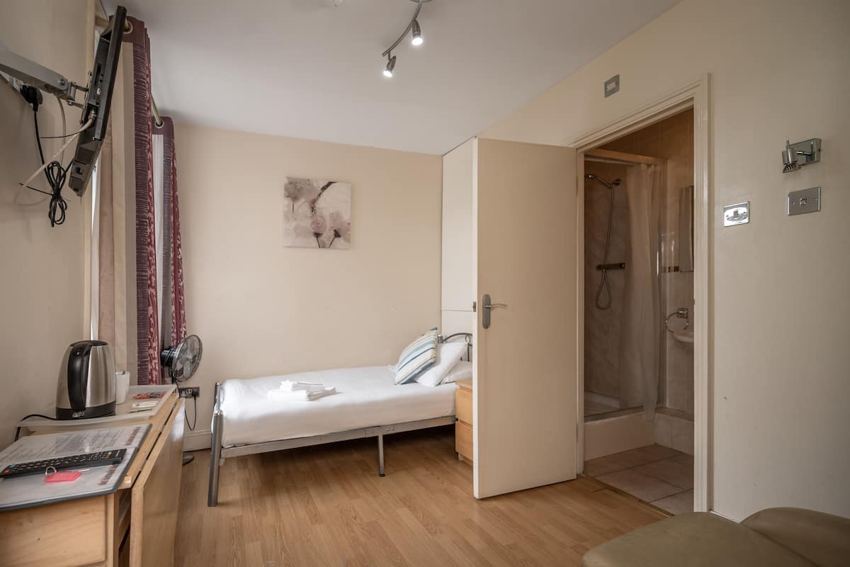 Una habitación pequeña y limpia con una cama individual, una mesita de noche con hervidor, un ventilador y zona de ducha.