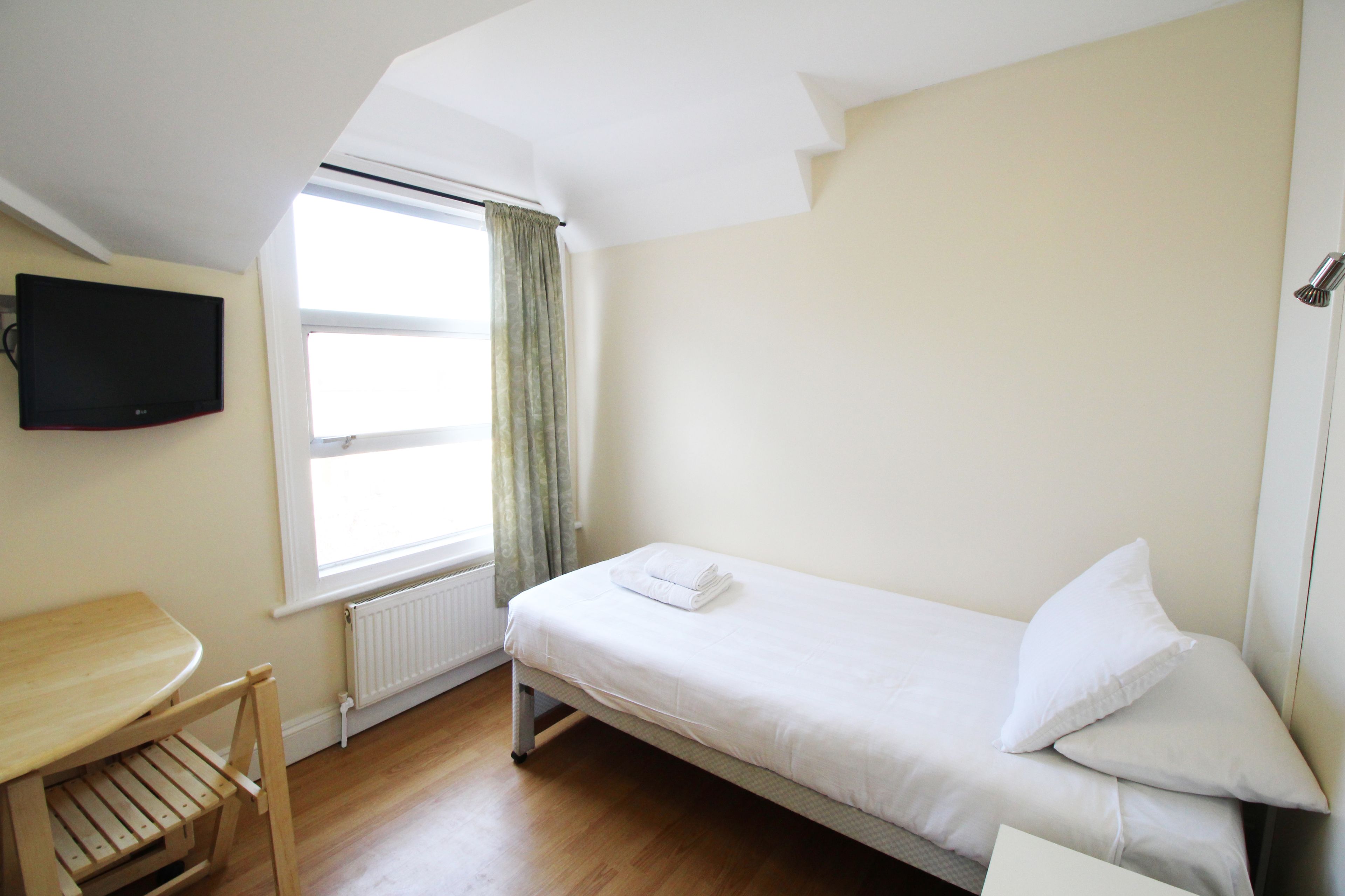 A small, single-bed room with a wooden desk and chair, a flat-screen TV mounted on the wall, and a window with curtains.