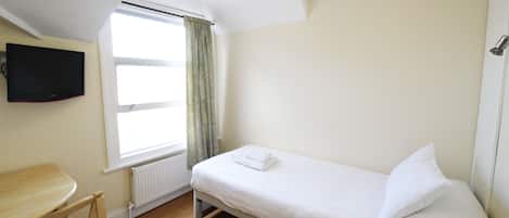 A small, single-bed room with a wooden desk and chair, a flat-screen TV mounted on the wall, and a window with curtains.