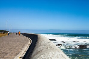 A coastal walkway with a concrete barrier, ocean waves, and a clear blue sky.