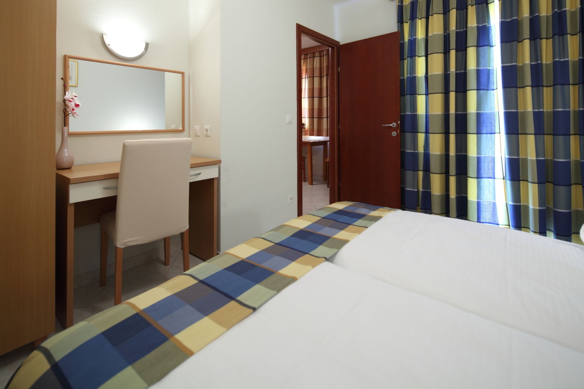A hotel room with a bed, a desk, and a chair. There is a door leading to another room.