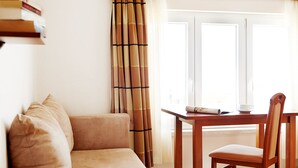 A cozy room with a sofa, a wooden table, and a chair. There is a window with curtains, and a shelf with books and a cup. - Apartments Ivana (Baska Voda)