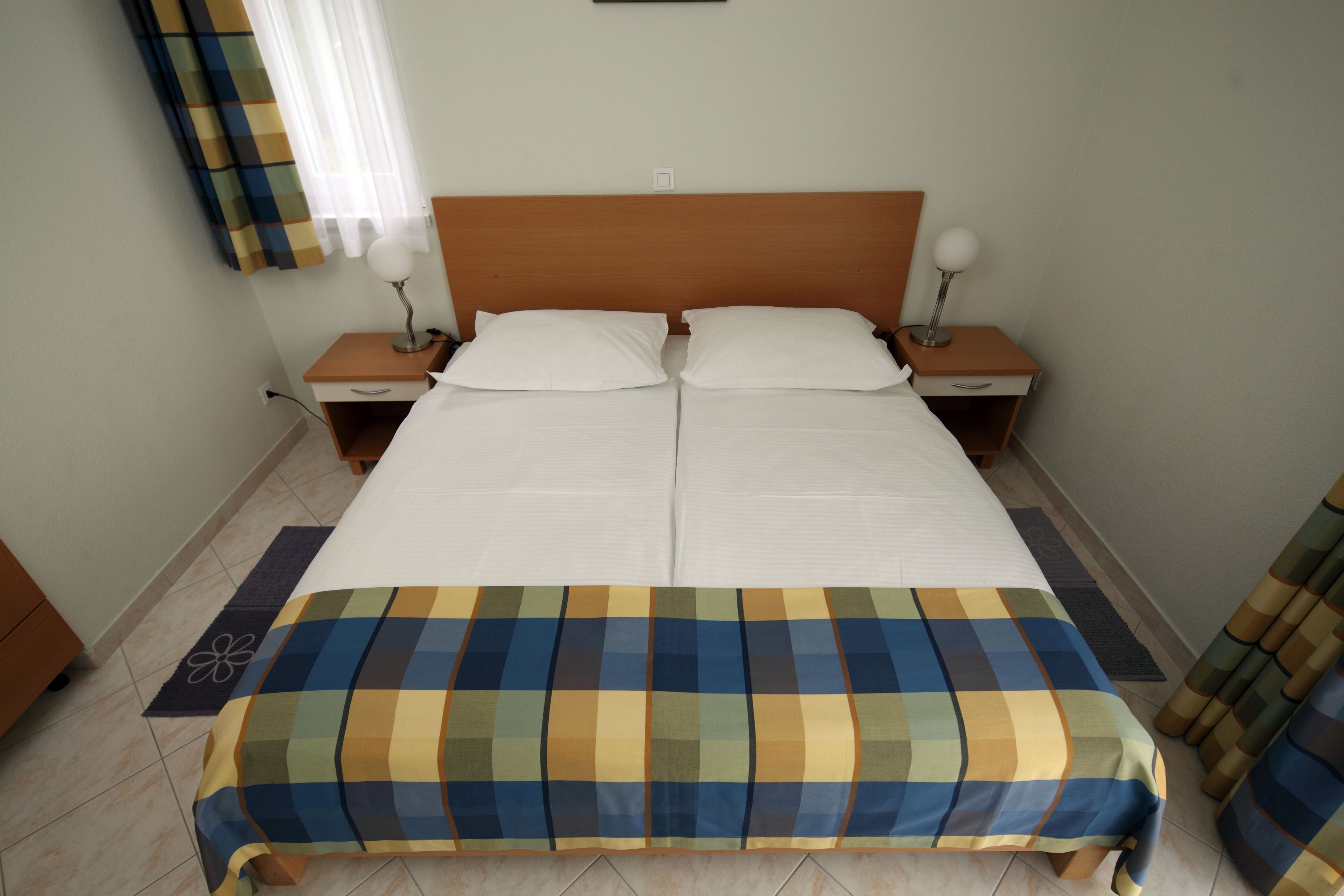 Una cama doble con colcha a cuadros, dos mesitas de noche con lámparas, una mesilla y una ventana con cortinas.