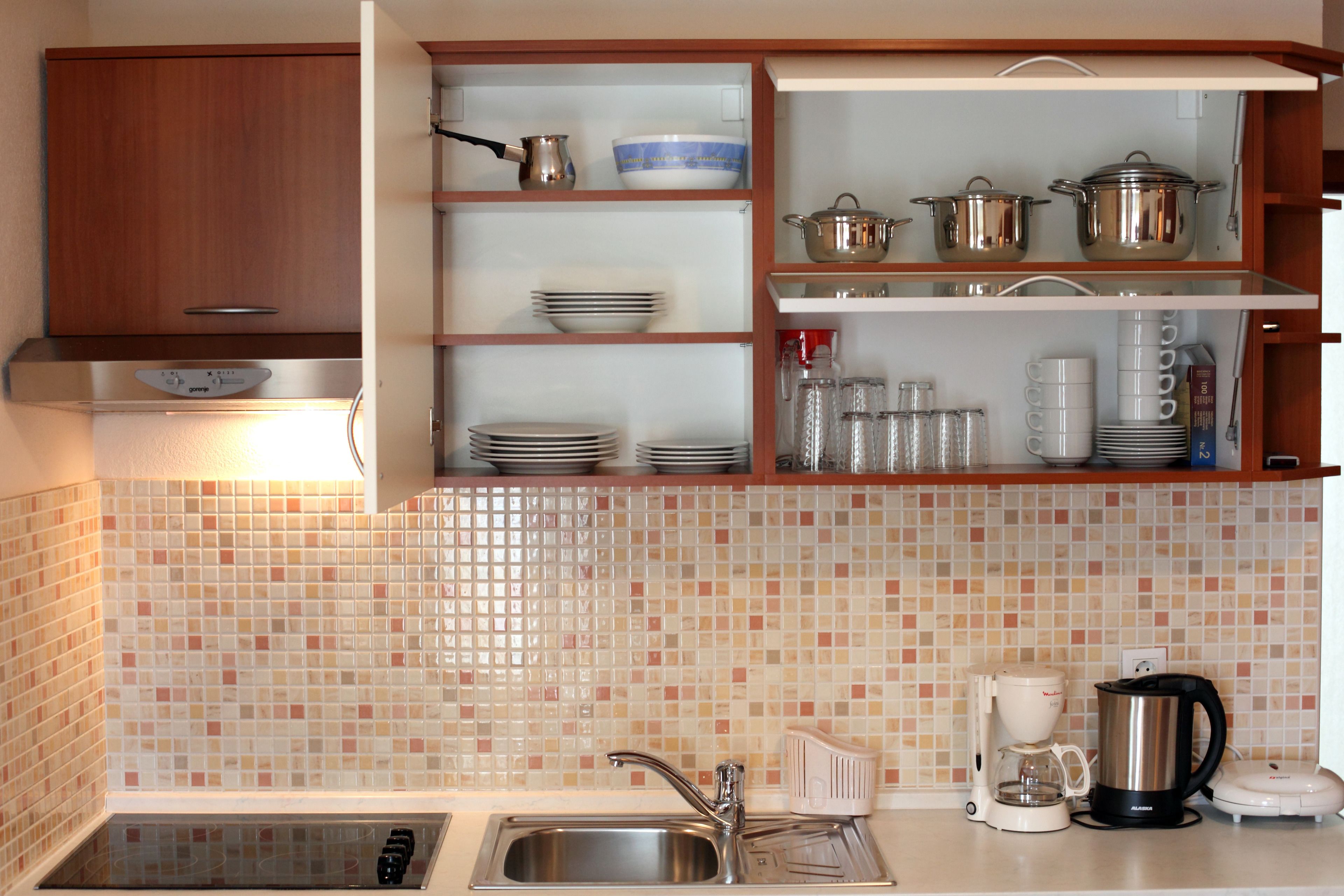 Una cocina con un lavamanos de acero inoxidable, una cafetera y un hervidor. Hay estantes con vajilla y un microondas.