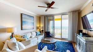 A spacious living room with a large sofa, armchairs, a coffee table, and a TV. A ceiling fan is visible, and the room has a large window with a view of the ocean. - Azure Condominiums (Fort Walton Beach)