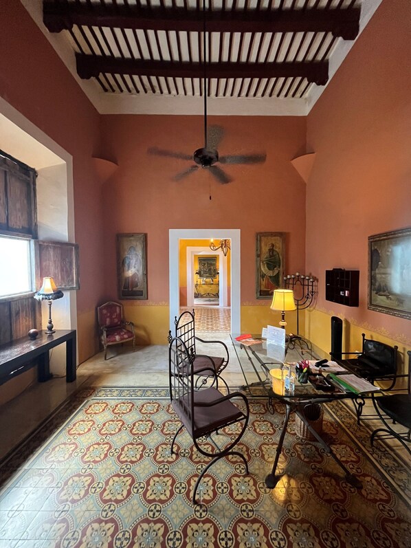 A room with a patterned floor, a ceiling fan, a desk with a chair, a table with a lamp, and a chair with a cushion.
