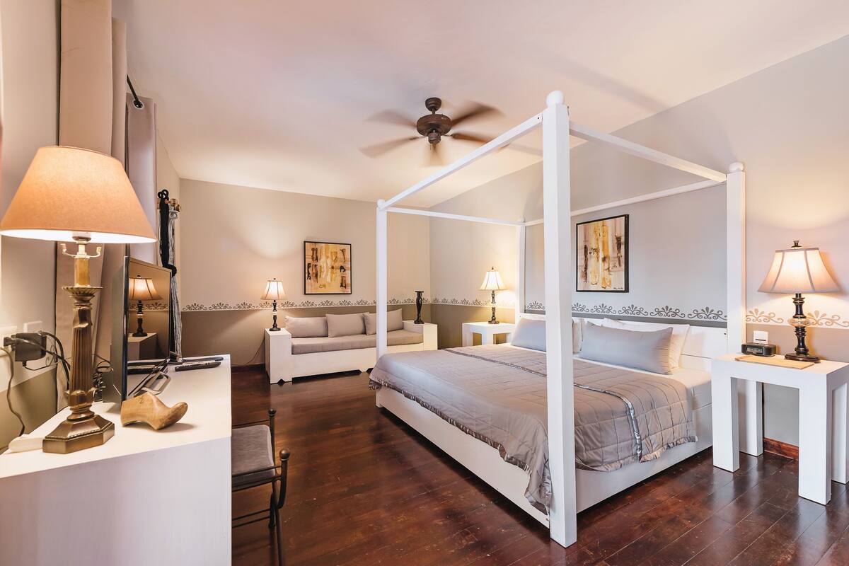 A four-poster bed with a canopy, a bedside table with a lamp, a nightstand with a lamp, a sofa, and a chair in a bedroom with wooden flooring and a ceiling fan.