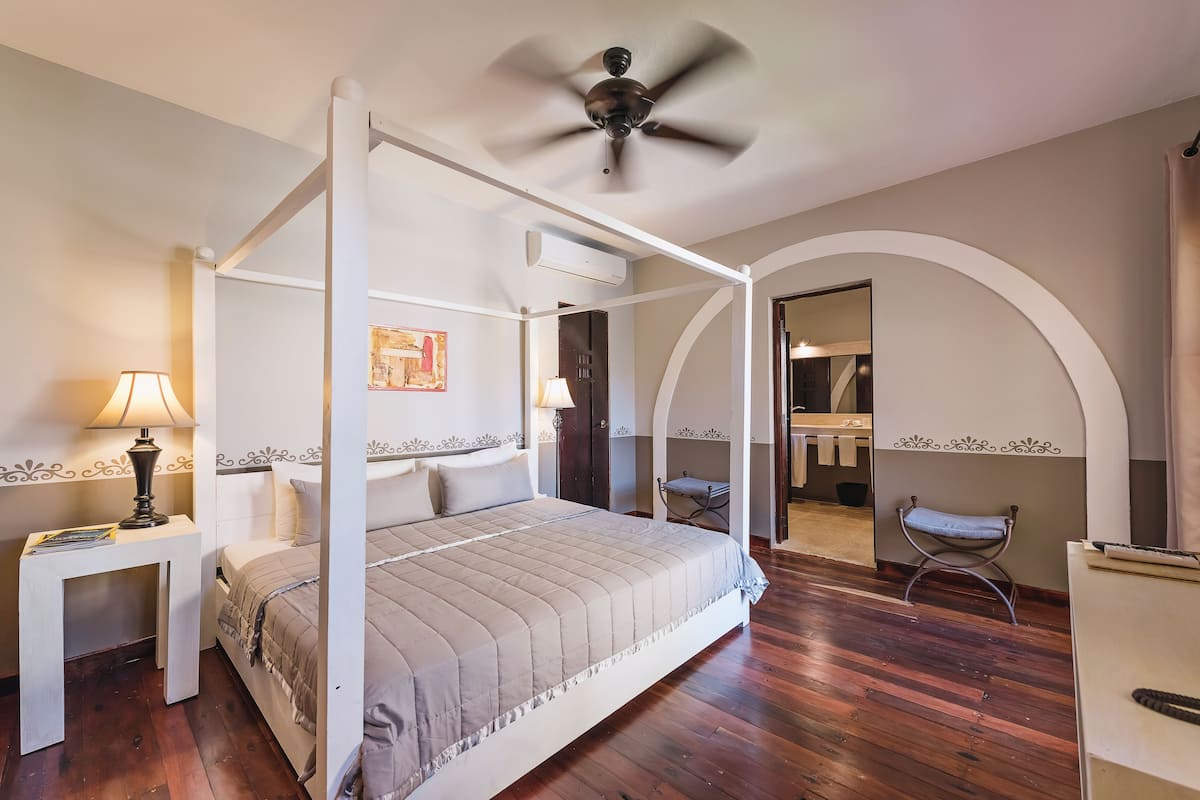 A four-poster bed with a ceiling fan, a bedside table with a lamp, and a wooden floor.