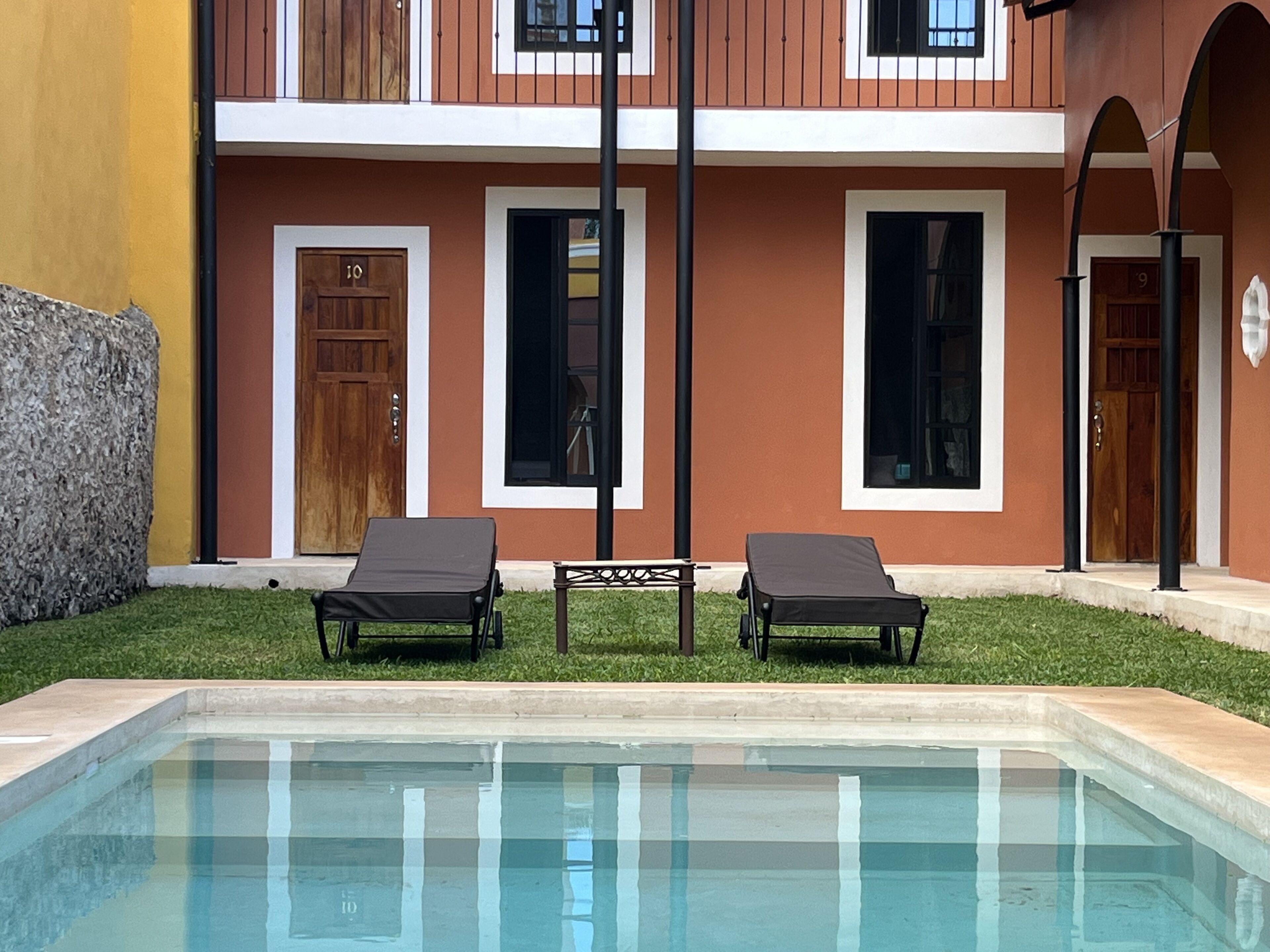 Area piscina con due poltrone da relax e un tavolino, circondata da un edificio con diverse porte e finestre.
