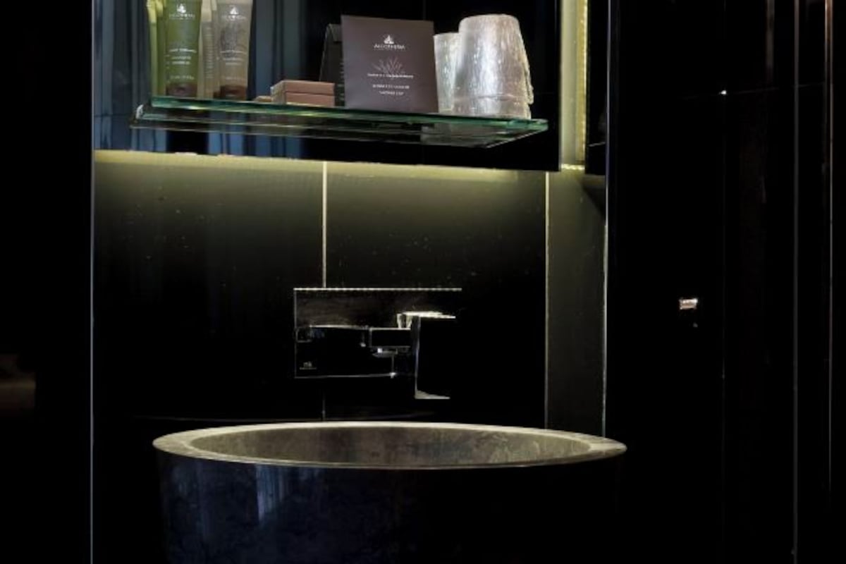 A modern bathroom with a large sink, a glass shelf with toiletries, and a dark wall.
