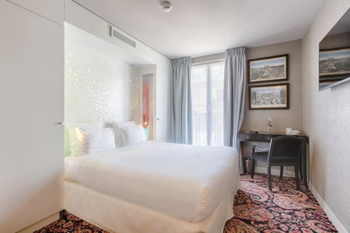 A hotel room with a large bed, a desk, and a chair. There are two windows with curtains, and the room has a patterned carpet.