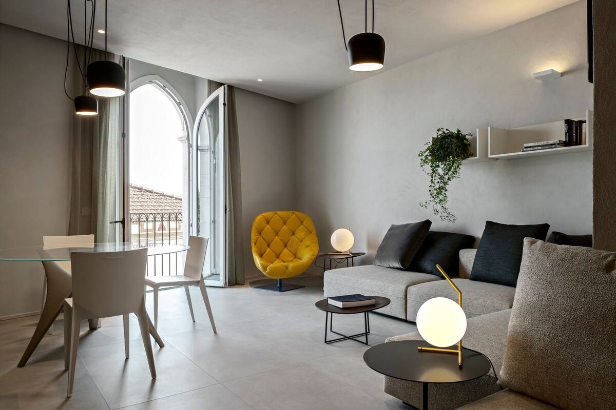 Een moderne woonkamer met een glazen eettafel, een gele fauteuil, een bank en een salontafel. Er zijn hanglampen, een plant en een boekenkast.